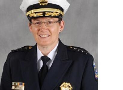 White woman smiling with wire-rimmed glasses wearing a large white police hat and a blue coat with a gold badge