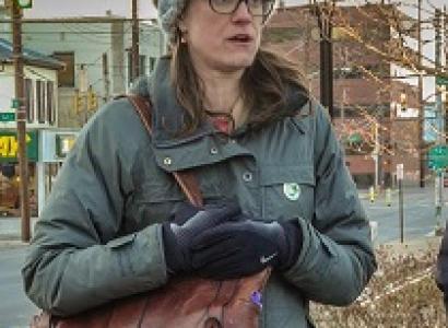 Young woman with brown rimmed glasses, long brown hair in winter clothes outside with her purse over her shoulder an holding it close to her body, looking like she is talking