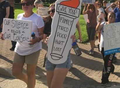 People marching outside in shorts with trees in background carrying signs, one saying Give us your tired poor
