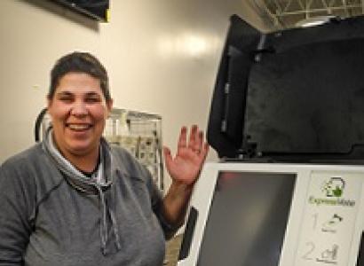 White woman with short brown hair smiling and displaying a white computer-looking machine with a big screen