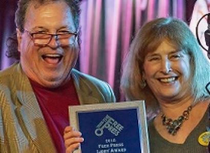 White man with glasses laughing posing with  white women with shoulder length blonde hair holding a plaque and smiling