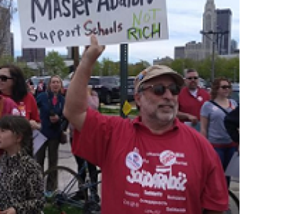 White man with sunglasses, a gray beard and a baseball cap wearing a Solidarity T-shirt is outside among lots of people with downtown Columbus in the background, holding a sign that says City Council Master Abators Support Schools not the Rich