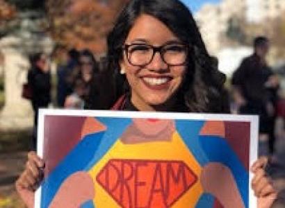 Young smiling woman with long black hair and big black-rimmed glasses holding a bright colorful sign that says DREAMER on it