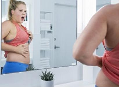 A blond woman with her hair in a knot of top of her head looking out as an image in a mirror with her mouth open and showing her belly between a red bra top and blue bottom with her hand on her belly