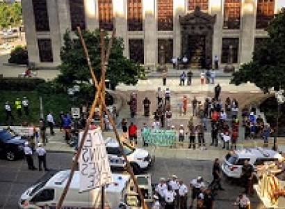 Scene in downtown with tall office buildings lots of people in the streets with protest signs, a lot of police and a tepee looking wooden structure