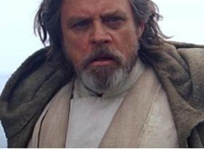 Older man with unkempt gray long hair and a beard looking intensely at the camera wearing a cape