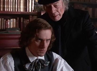 A man with chin length curly brown hair looking intense sitting down with old fashioned clothes from the 30s and an older gray haired man in black with a black top hat leaning over his shoulder saying something in his ear, bookshelves in the background