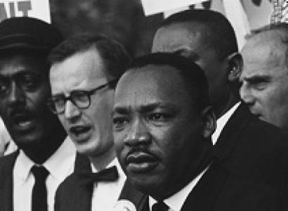 Black man in foreground, Martin Luther King Jr. surrounded by other men with sign above saying Now