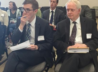 Two men in foreground of people sitting in chairs, one younger white man with dark rimmed glasses with his hand over his mouth holding a pen, and the other a white haired white man, both in dark suits