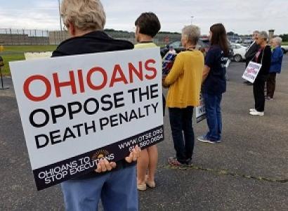 The backs of four people at a vigil outside one holding a sign that says Ohioans Oppose the Death Penalty