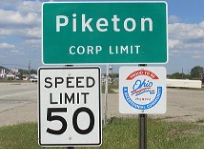 Green road sign saying Piketon Corp Limit, another sign saying Speed Limit 50 and a sign with an Ohio logo on it and in the background a highway and some grass and the blue sky with white clouds