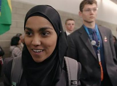 Young dark skinned woman in a burqa smiling and a young white man in a suit looking serious behind her