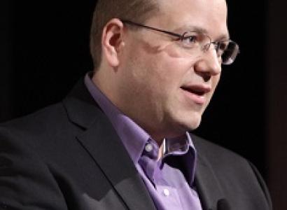 Middle aged white man with brown receding hair with wire rimmed glasses looking to the right with  a gray suitcoat and purple shirt his mouth open as if talking