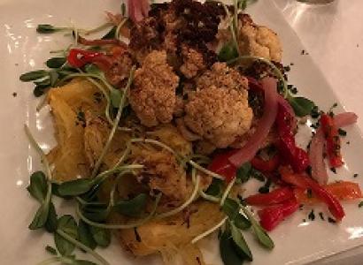 A white square plate on a table filled with greens, little toasty looking brownish balls, squash and red slivers of food