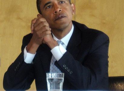 Obama with elbows on table clasping his hands together looking pensive