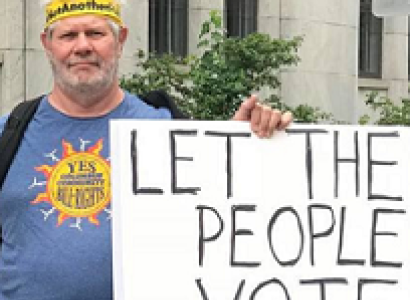 White man with yellow headband and blue T-Shirt with sun in the middle holding a sign that says Let the People Vote