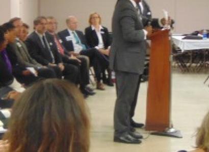 Lots of men in suits sitting in a row on chairs facing right and two black men in suits standing at a podium
