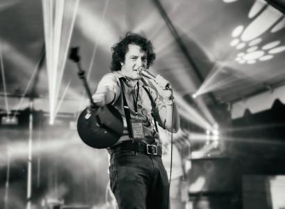 Black and white photo of young dark haired man singing into a mic on a stage
