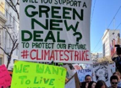 Outdoor rally with signs, one says We Want to Live Here, one says Dear Nancy Pelosi will you support a Green New Deal