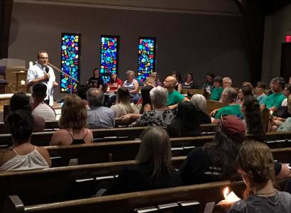 Man in white shirt talking into a mic at the far left side and a congregation of people sitting in pews and three stained glass windows in the back