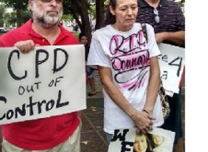 Man with gray beard and sunglasses wearing a baseball cap holding a sign that says CPD out of control standing next to an older woman with gray hair pulled back in a ponytail looking down at the ground, very sad, wearing a white T-shirt with words "RIP Donna" on it and holding a sign with pictures of a young woman on it