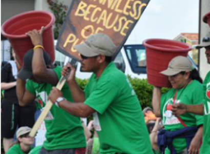 Dark-skinned men and women in green shirts at a demonstration marching to the left one holding a sign saying Penniless because of and two others holding big rust colored buckets