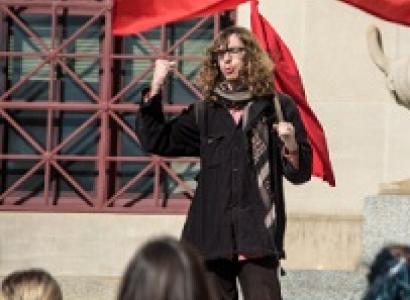 Young white man with long brown curly hair and glasses holding a very large red flag standing in front of a crowd raising his fist in the air
