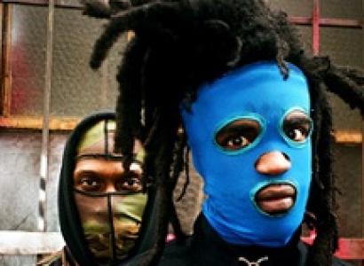 Two men one in front with dreds sticking up off his head and a bright blue mask and a guy behind him with a camouflage mask