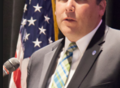 Middle aged white man in a suit with brown receding hair at a mic with a flag in the background