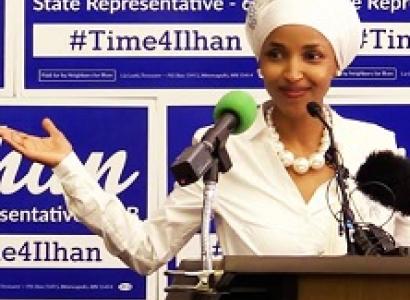 Young woman of color wearing a white hat and suit and pearl necklace standing in front of mics at a podium and signs with her name Ilhan State Representative in the background