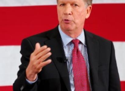 White man middle-aged in dark suit with red tie and white shirt talking and holding up his hand by his chest in front of a red and white striped background