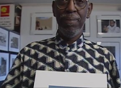 Older black man, bald with gray mustache and beard in a gray and white shirt holding a photo and standing in front of lots of other photos on the wall. 