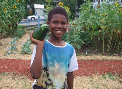 Young black boy smiling and holding a very large zucchini on his shoulder, standing in a garden