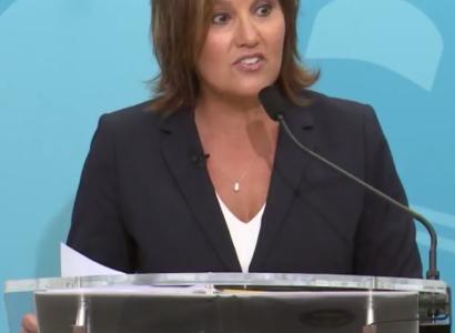 White woman in a black suit with white shirt underneath and necklace with shoulder length brown hair standing at a mic with a blue background, her mouth in a form like she's talking