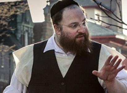 Large white man with jewish hat and wire rimmed glasses, a longish brown beard wearing a white collared button down shirt and black button vest his hand gesturing as he talks to someone to his right