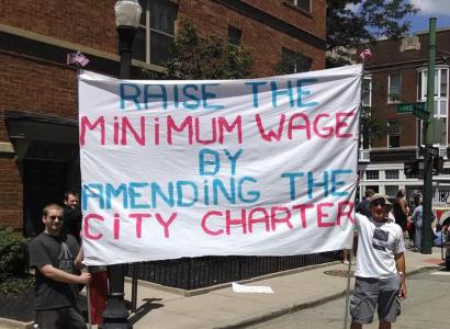 Two men holding sign reading Raise the MInimum wage by amending the city charter