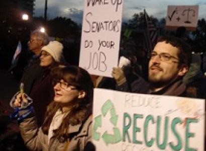 People standing outside in winter coats at dusk one white man with brown hair, facial hair and glasses with a sign that reads Recuse and a white woman next to him 
