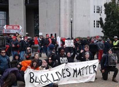 People wearing winter clothes bending down on one knee outside holding a long white banner with black letters reading Black Lives Matter in front of a large white building