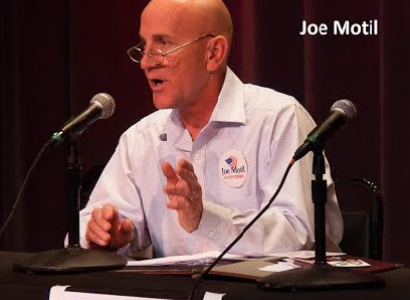 Bald white man with little wire-rimmed glasses talking at a mic on a table, wearing a white shirt and gesturing with his left hand