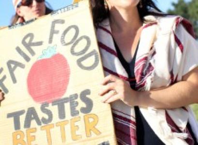White women with long dark hair and sunglasses holding sun that says Fair Food Taste Better