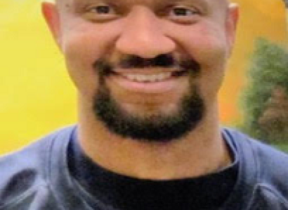 Bald black man with dark mustache and goatee, wearing blue shirt smiling