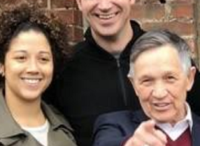 Young black woman smiling on the left, tall white man smiling in the middle and older white man pointing at the camera standing in front of a brick wall
