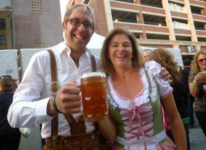 White ma with gray hair and glasses smiling holding a huge mug of beer with a woman who is wearing a German-looking corset and frilly top