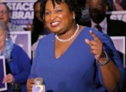 Older black woman with short curly black hair, white pearl necklace, blue dress, standing and smiling while gesturing in front of lots of people holding signs that say Stacey Abrams