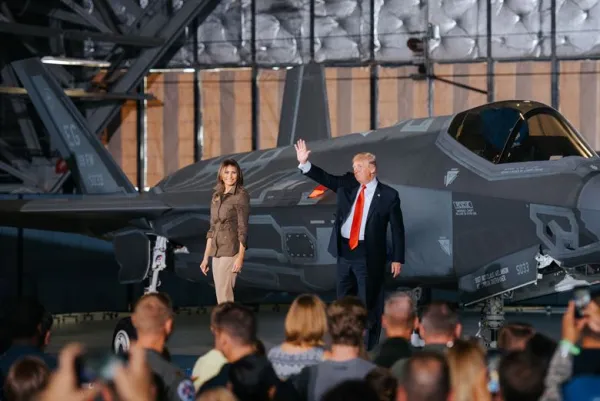 Trump, heavyset man in suit with red tie waving next to wife Melania, shapely woman with long brown hair standing in front of a military plane with people in foreground facing him