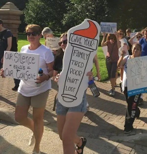 People marching outside in shorts with trees in background carrying signs, one saying Give us your tired poor