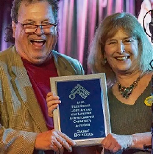 White man with glasses laughing posing with  white women with shoulder length blonde hair holding a plaque and smiling