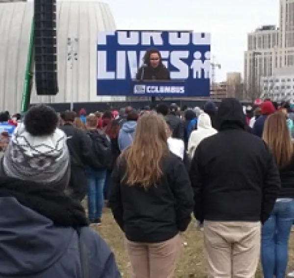 Backs of people standing watching a speaker on a screen