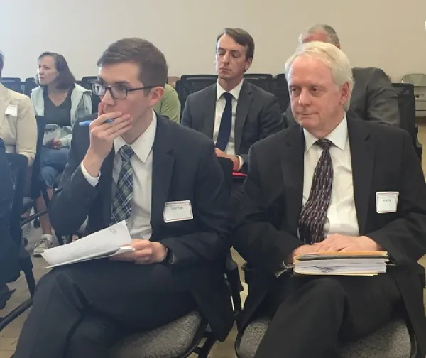 Two men in foreground of people sitting in chairs, one younger white man with dark rimmed glasses with his hand over his mouth holding a pen, and the other a white haired white man, both in dark suits