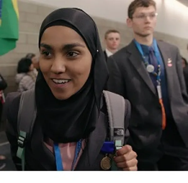 Young dark skinned woman in a burqa smiling and a young white man in a suit looking serious behind her